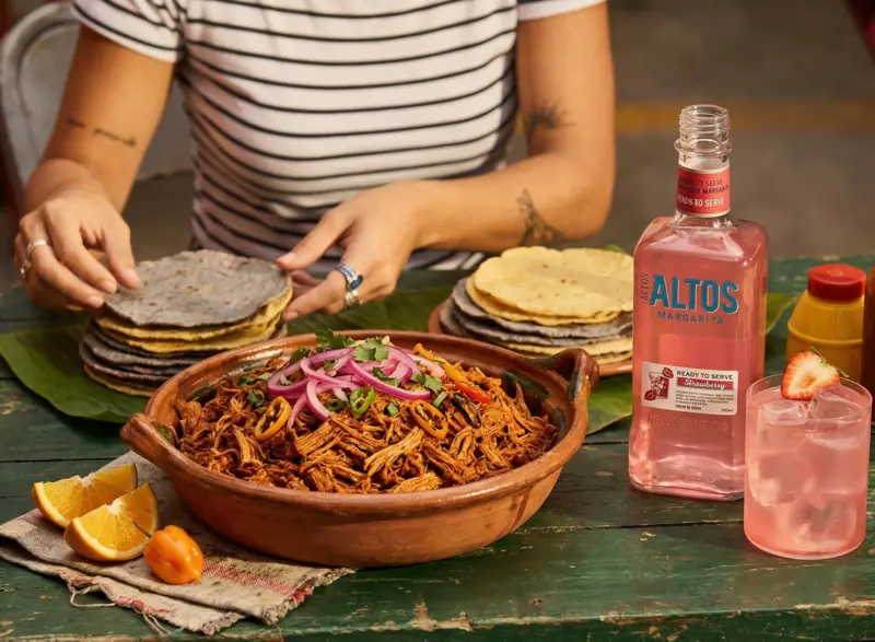 botella de altos strawberry margarita junto a cochinita pibil, tortillas y cebolla morada encurtida sobre una mesa rustica