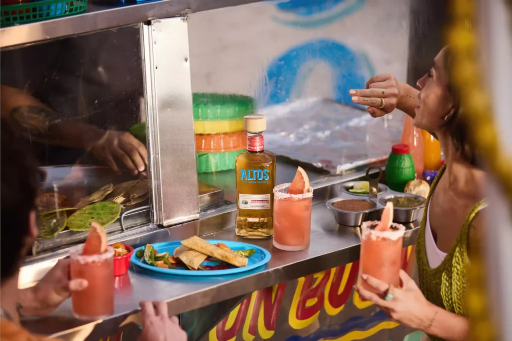 A bottle of Altos Reposado tequila on a bar counter beside a Tequila Sour served over a large ice cube in a rocks glass.
