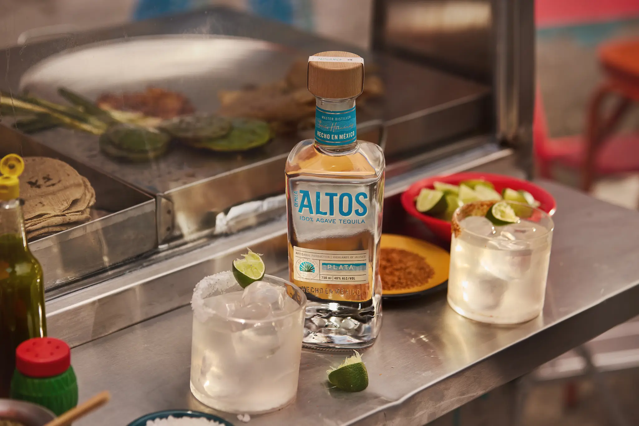  A bottle of Altos Plata tequila and two skinny margaritas on the rocks sitting on a stainless steel food cart counter, with tacos cooking on a grill in the background. 