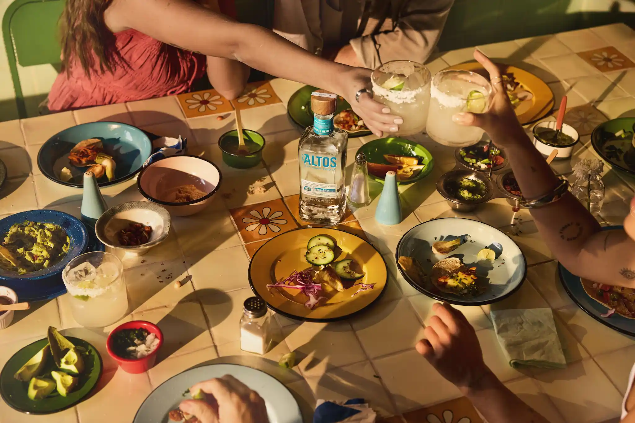Vista cenital de amigos brindando con margaritas sobre una mesa de azulejos iluminada por el sol, llena de platos con tacos, guacamole y una botella de tequila Altos Plata.