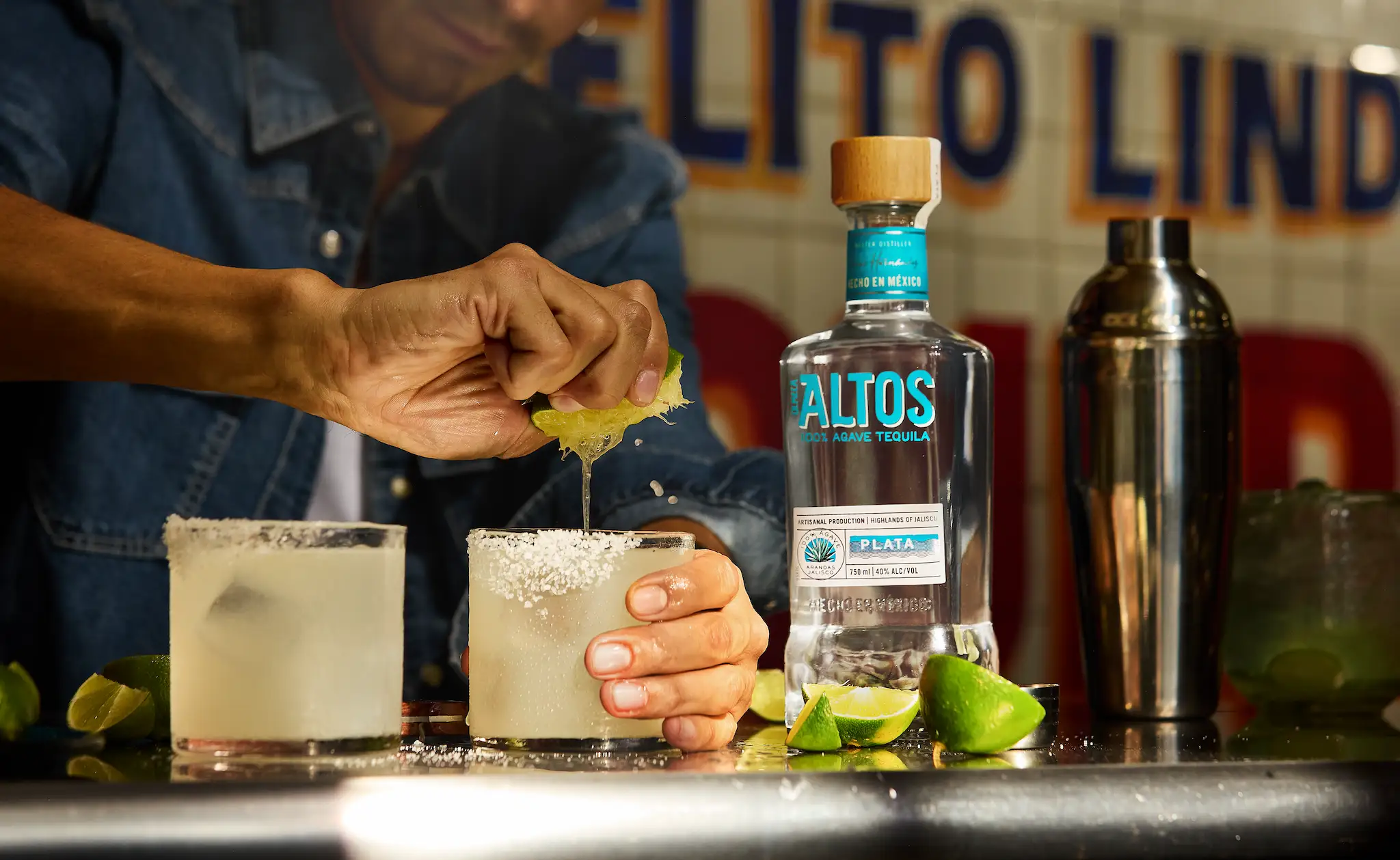  Close-up of a person squeezing a fresh lime into a salt-rimmed glass to make a skinny margarita, next to a bottle of Altos Plata tequila and a cocktail shaker. 