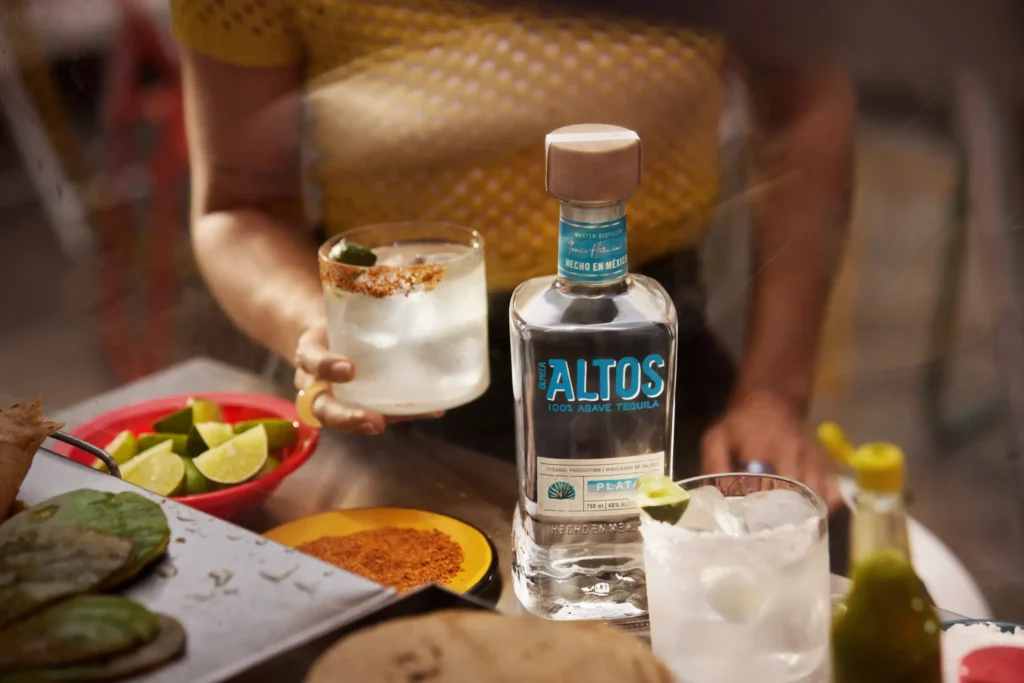 A bottle of Altos Plata tequila in sharp focus on a metal counter, with a person holding a margarita in the blurred background. 
