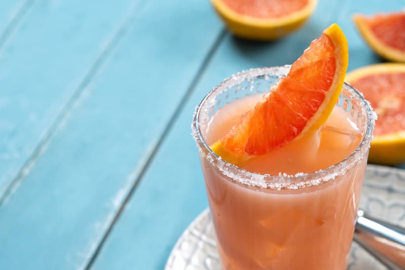 Cóctel Paloma clásico preparado con tequila Olmeca, servido en vaso escarchado con sal y rodaja de toronja fresca sobre superficie de madera turquesa