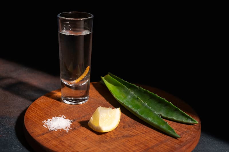 Bebida de agave servida en vaso con limón, sal y hojas frescas de agave sobre tabla de madera
