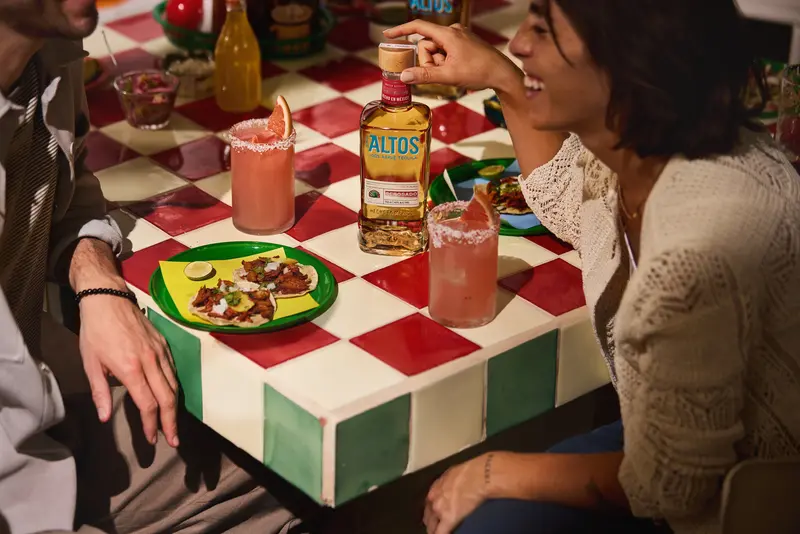 bottle of altos reposado on a table with pink cocktails and mexican food plates
