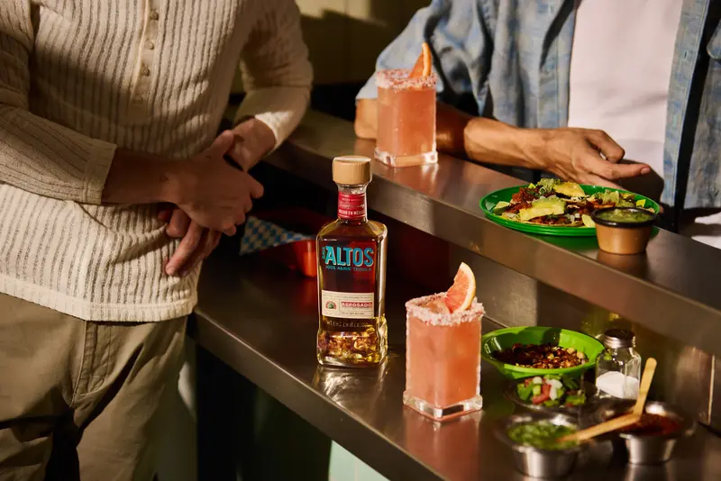 bottle of olmeca altos reposado tequila on a counter with pink cocktails and mexican food