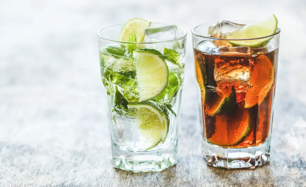 two tall glasses with ice and lime, one with a clear soda and mint and one with tequila and cola