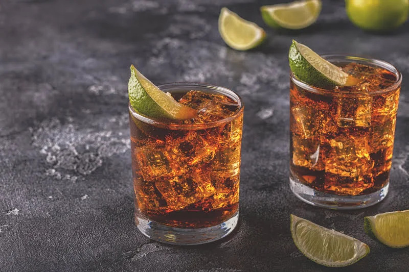 two short glasses of tequila and cola with ice and lime wedges on a dark background