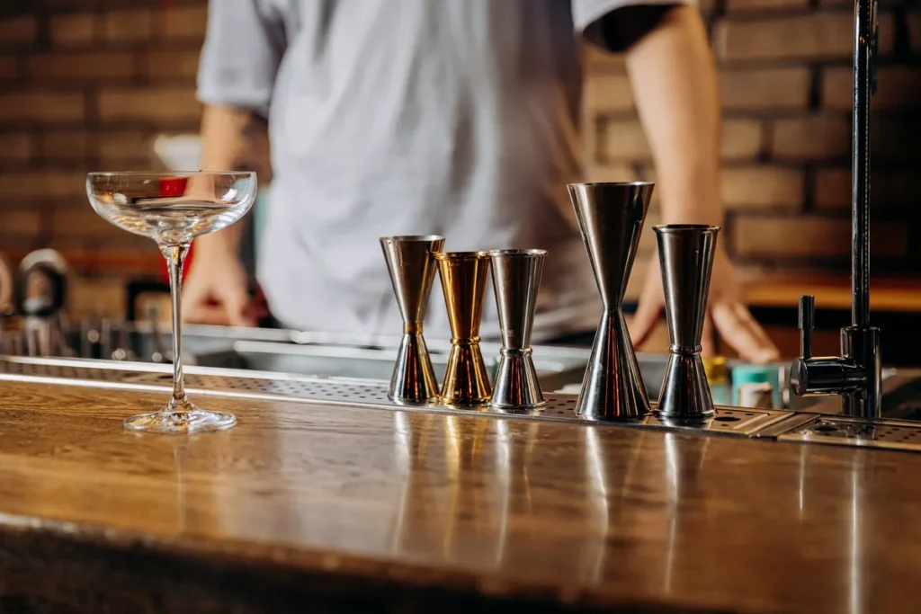 different metal cocktail jiggers lined up on a bar next to an empty coupe glass.