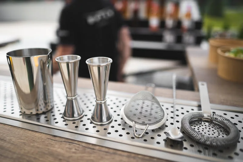 herramientas de coctelería de acero inoxidable sobre una barra, incluyendo jiggers, una coctelera, un colador y una cuchara de bar.
