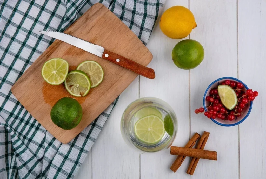 Top view of a wooden board with sliced limes, a knife, a glass of water with lime slices, a lemon, red berries, and cinnamon sticks on a white table.