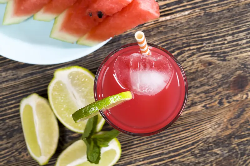 top view of a pink watermelon cocktail with ice, lime, and fresh watermelon slices on a wooden table.


