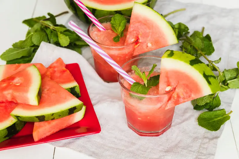 two fresh watermelon drinks with mint and striped straws, served with watermelon slices on a table.