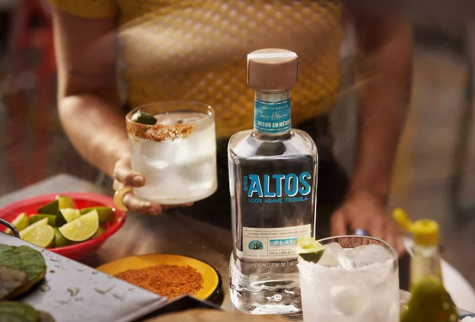 Preparación de un cóctel Paloma con tequila Olmeca Altos Plata, con gajos de lima fresca, vaso con borde de sal y la botella de Altos como protagonista de la escena.