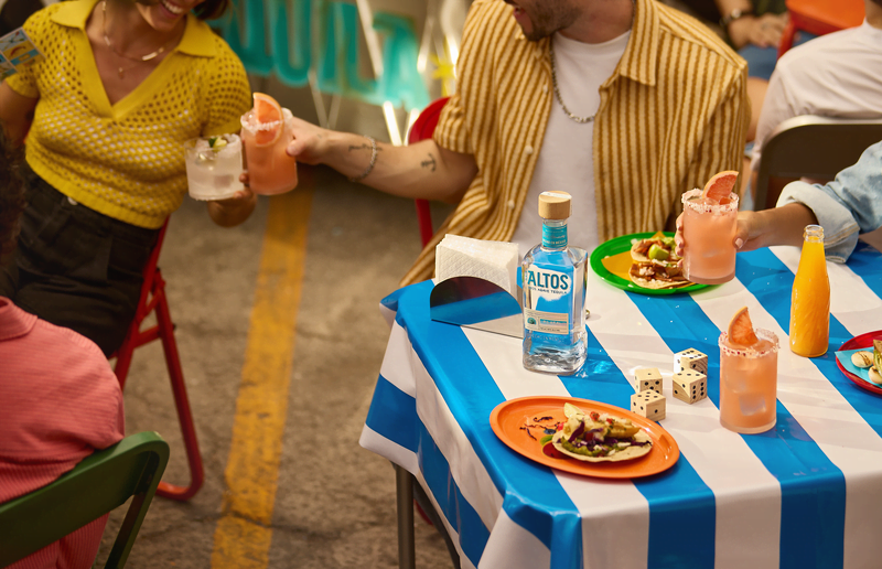 Olmeca Altos tequila bottle on a blue-and-white table as people toast pink grapefruit Palomas alongside tacos.