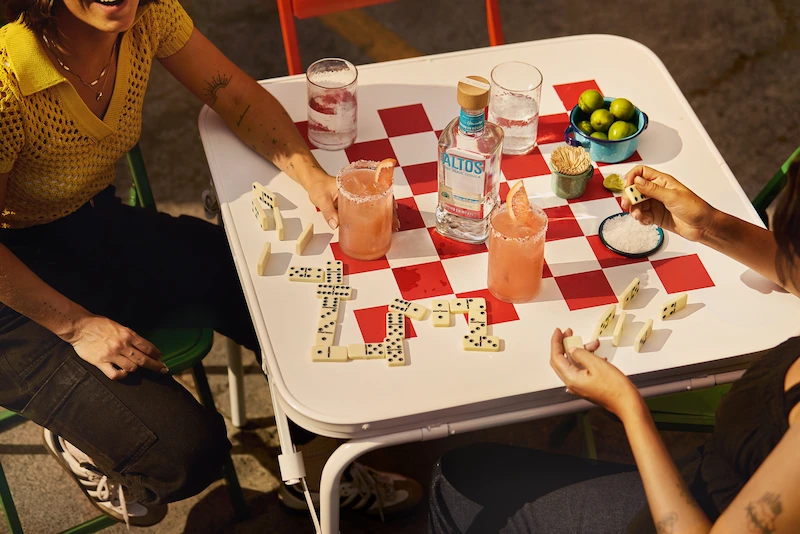 two people enjoying pink margaritas at a table with dominoes, limes, and a bottle of olmeca altos tequila.