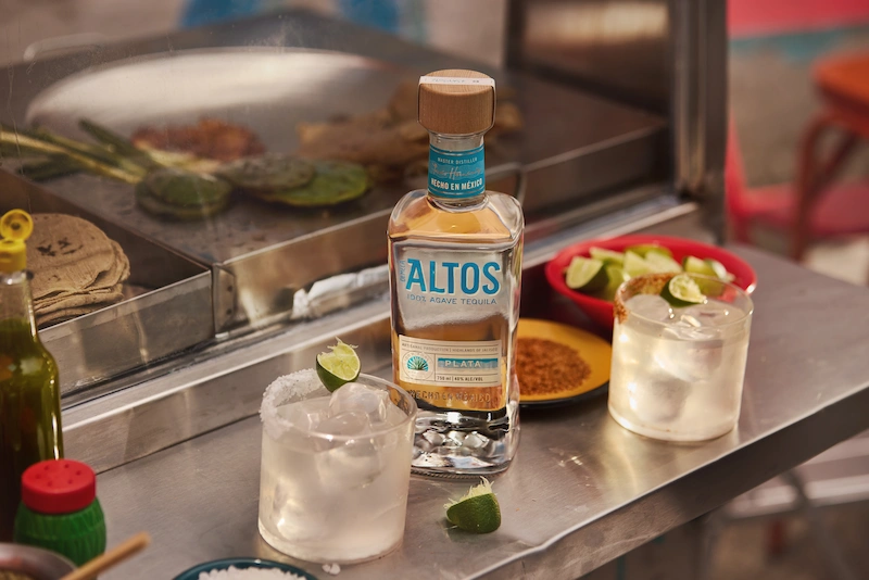 bottle of olmeca altos plata tequila with two margaritas in rocks glasses, served with lime wedges on a metal bar counter.