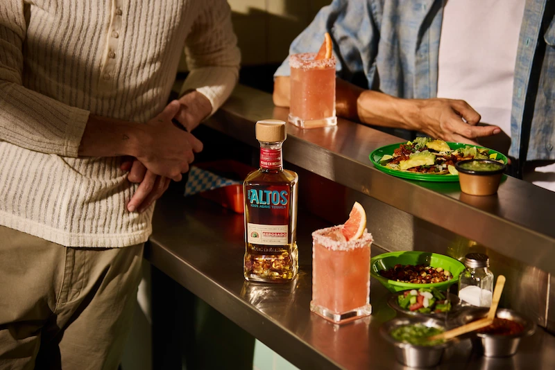 bottle of altos reposado tequila with grapefruit cocktails and food plates on a bar counter

